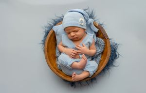 A sleeping newborn wearing light blue sleepwear and hat rests in a round wooden basket with gray fur bedding.