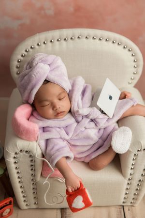 A plush armchair holds a sleeping baby in cozy lavender bathrobe and slippers holding a white device.