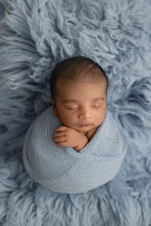 A sleeping newborn wrapped in a soft blue knit blanket lies on fluffy blue fur backdrop during professional photo session.