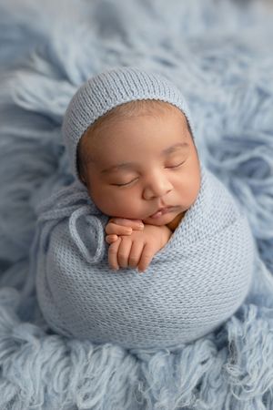 Sleeping newborn wrapped in light blue knitted swaddle and hat resting on soft blue textured blanket.