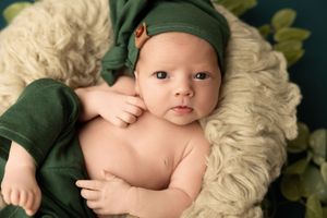 Baby wearing green bonnet rests on cream-colored fur blanket during professional portrait session.