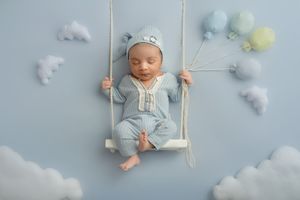 Sleeping baby in blue onesie on wooden swing with decorative clouds in blue background.