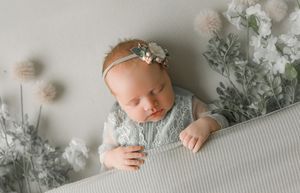 A sleeping baby in a lace outfit lies on a white textured surface with dreamy silver floral decor and dandelions.
