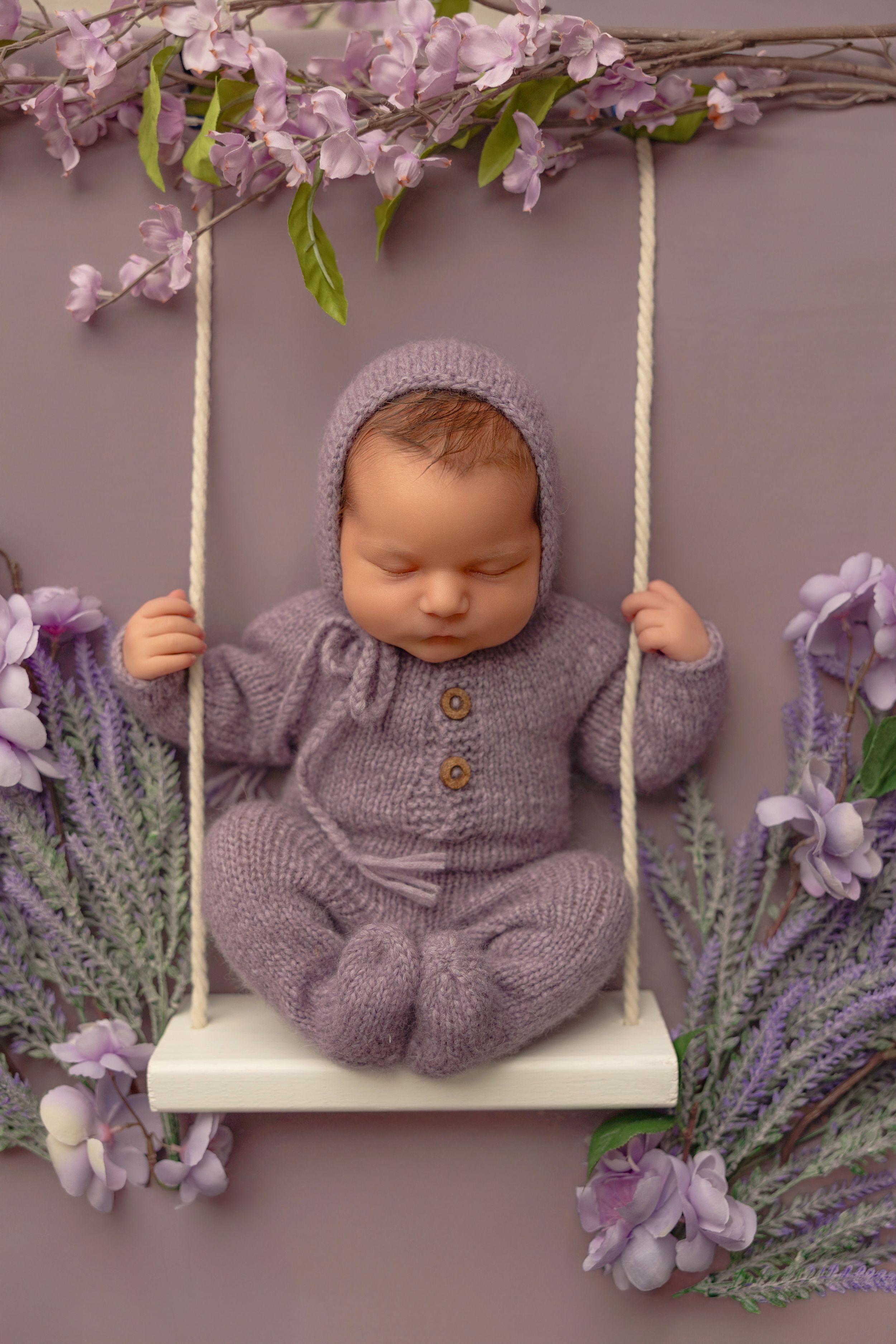 Newborn baby wearing a cozy purple knit outfit sleeps peacefully on a white swing surrounded by lavender flowers.