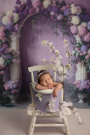 Sleeping newborn rests peacefully on white chair surrounded by purple and white floral arrangements in dreamy setting.