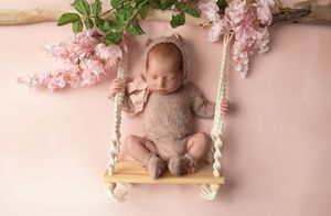 A peaceful sleeping baby in a knit romper rests on a wooden swing beneath delicate pink cherry blossoms.