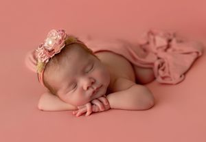 Sleeping newborn wearing pink floral headband on soft coral backdrop with matching fabric.