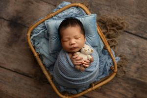 Sleeping newborn wrapped in soft blue swaddle blanket rests peacefully in woven basket on wooden floor.