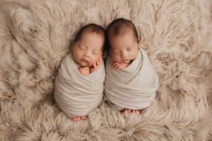 Two sleeping newborn twins swaddled in gray wraps lying on soft beige furry blanket.