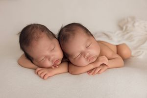 Two sleeping newborn babies lying side by side on soft white fabric in a peaceful pose.