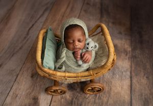 A sleeping baby wearing a mint green outfit lies in a wicker bassinet with wheels on wooden floor.