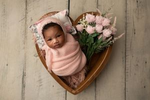 A sleeping newborn swaddled in soft pink fabric lies in a heart-shaped wooden bowl beside pink flowers.
