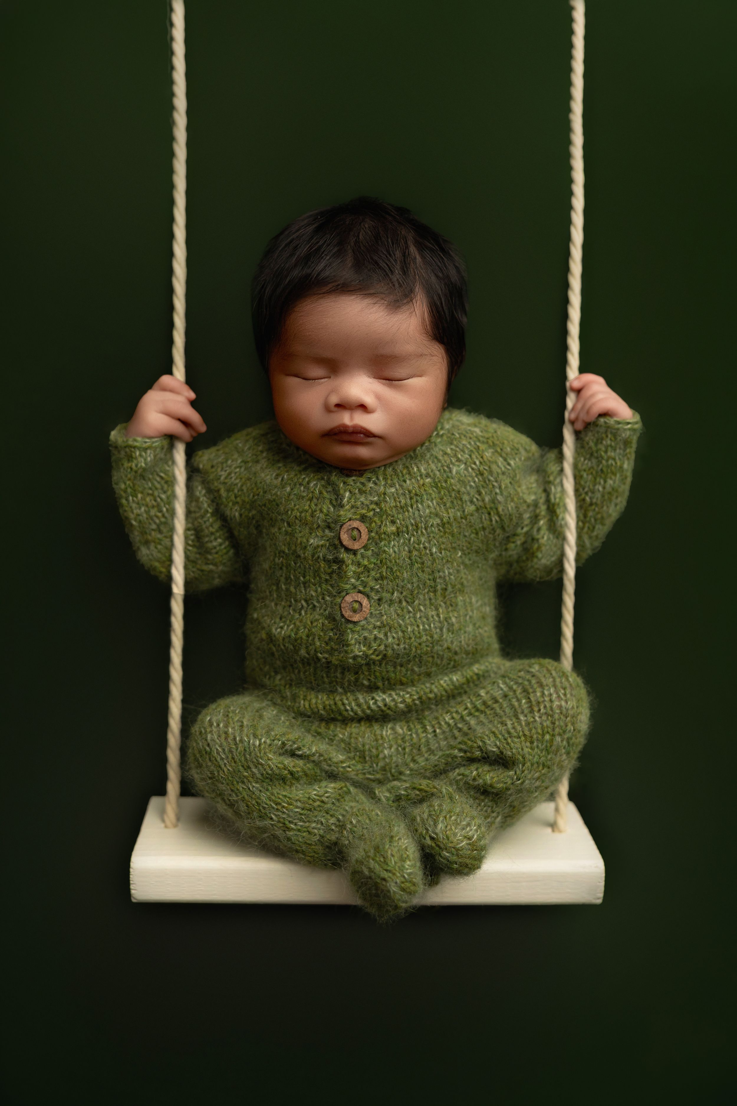 Newborn baby in green knit romper sleeping peacefully on a white swing with rope against dark green background.