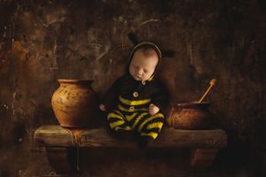 A baby dressed in a bee costume sits on a rustic wooden table with clay pots in a dark moody setting.