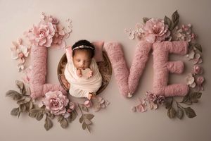 A newborn baby nestled in pink LOVE letters decorated with roses and floral arrangements for a creative photography setup.