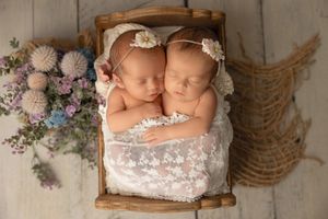 Two sleeping newborn babies rest in a wooden basket with lace blanket and lavender floral decorations.