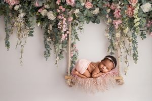 Newborn baby sleeping peacefully on pink swing beneath cascading floral arch with roses and eucalyptus.