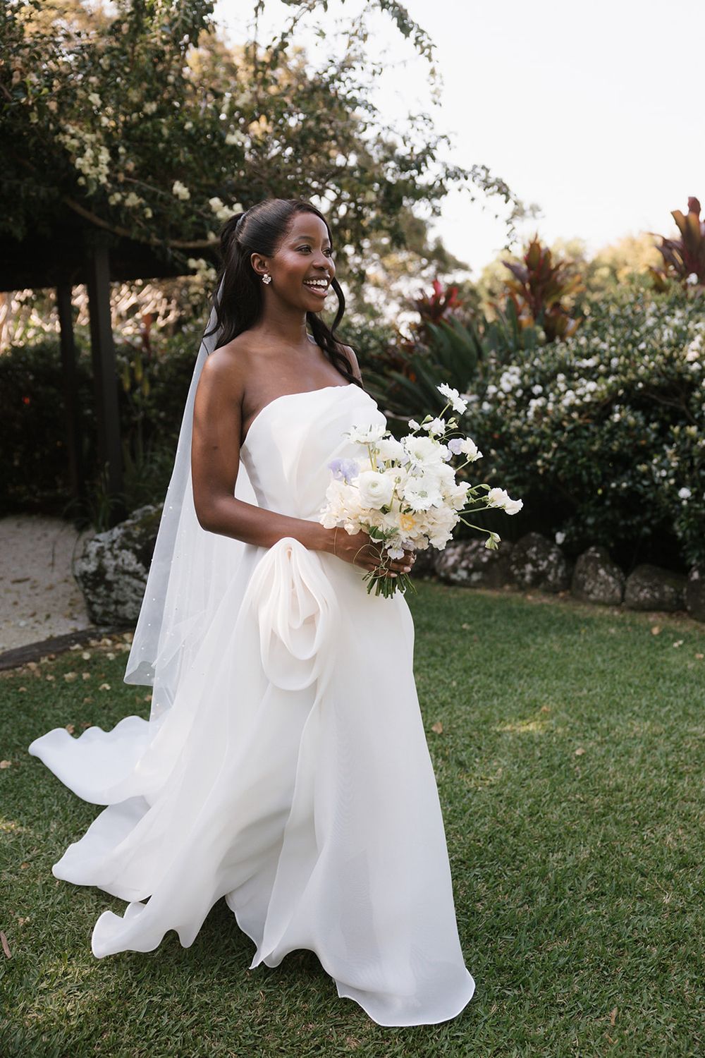 A joyful bride in a flowing white strapless gown holds a delicate bouquet of white flowers in a garden setting at sunset.