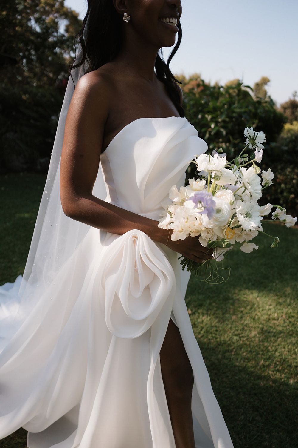 A bride in a flowing white strapless gown holds an elegant bouquet of white flowers on a sunny outdoor lawn.