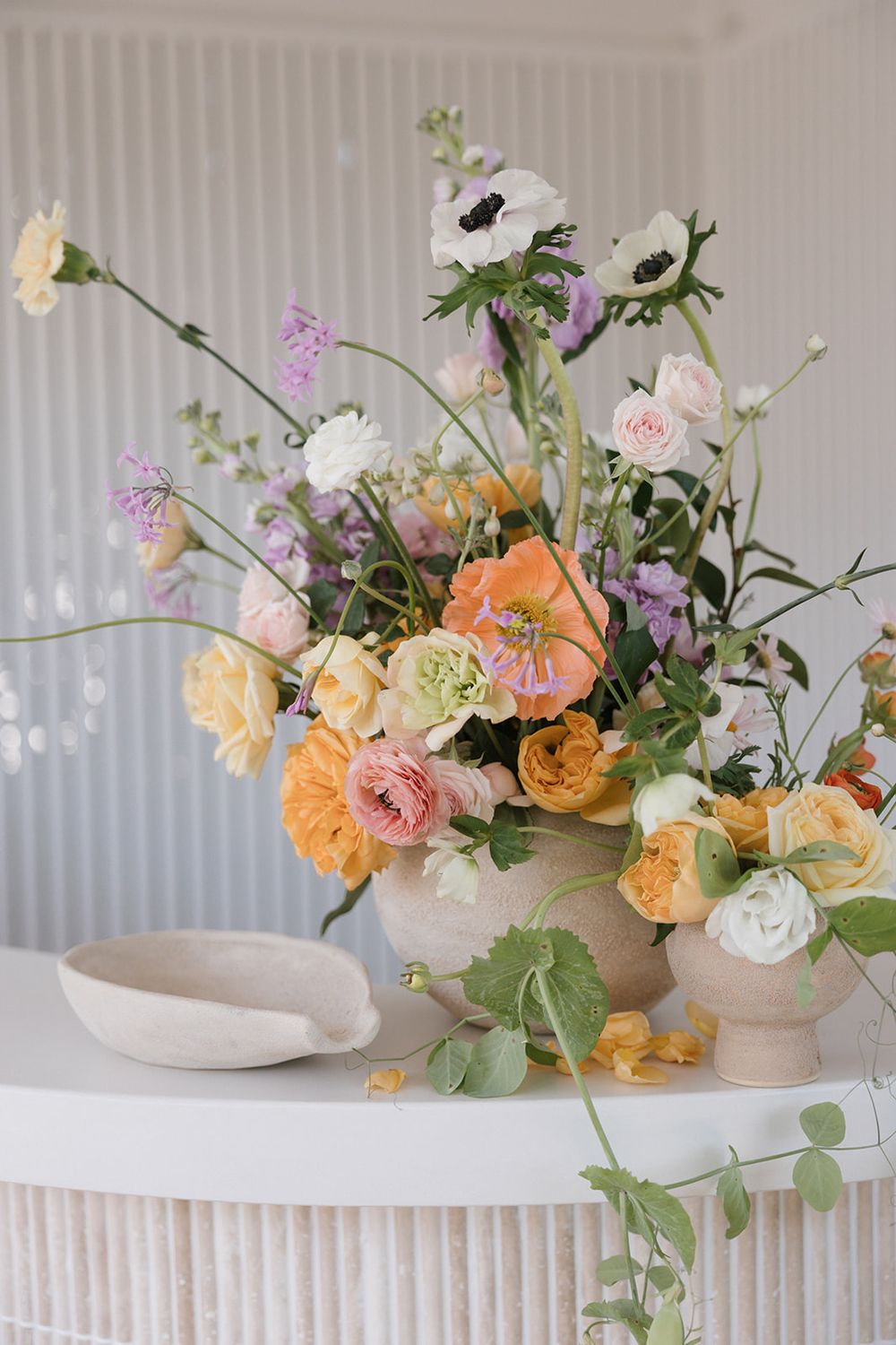 A romantic floral arrangement with peach, white, and purple blooms in a stone vase on a textured white surface.