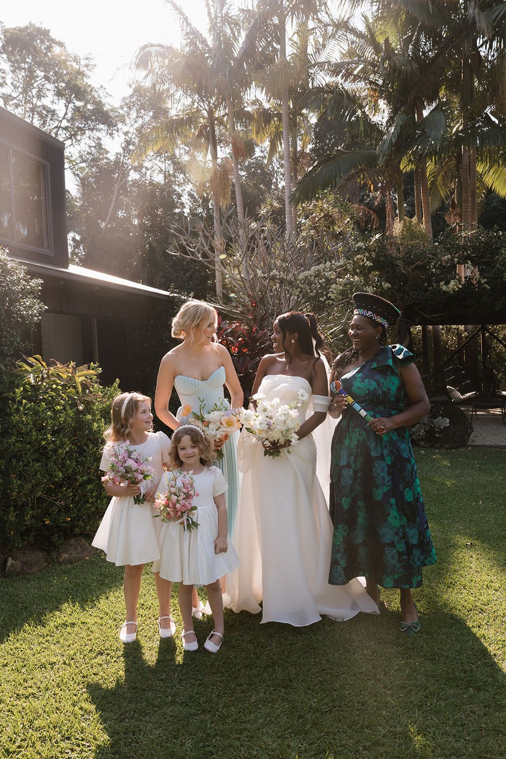 Wedding party including flower girls and bridesmaids posing together outdoors with palm trees in background.