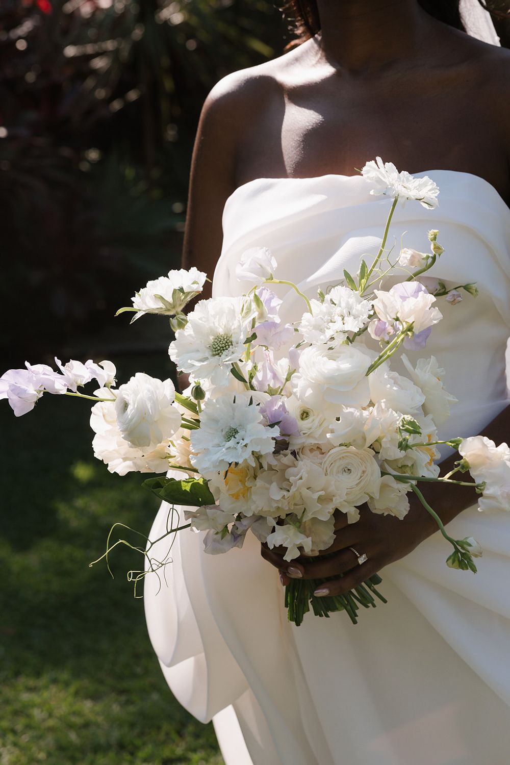 A delicate white wedding bouquet with cosmos and mixed flowers against a flowing white wedding dress in sunlit garden.