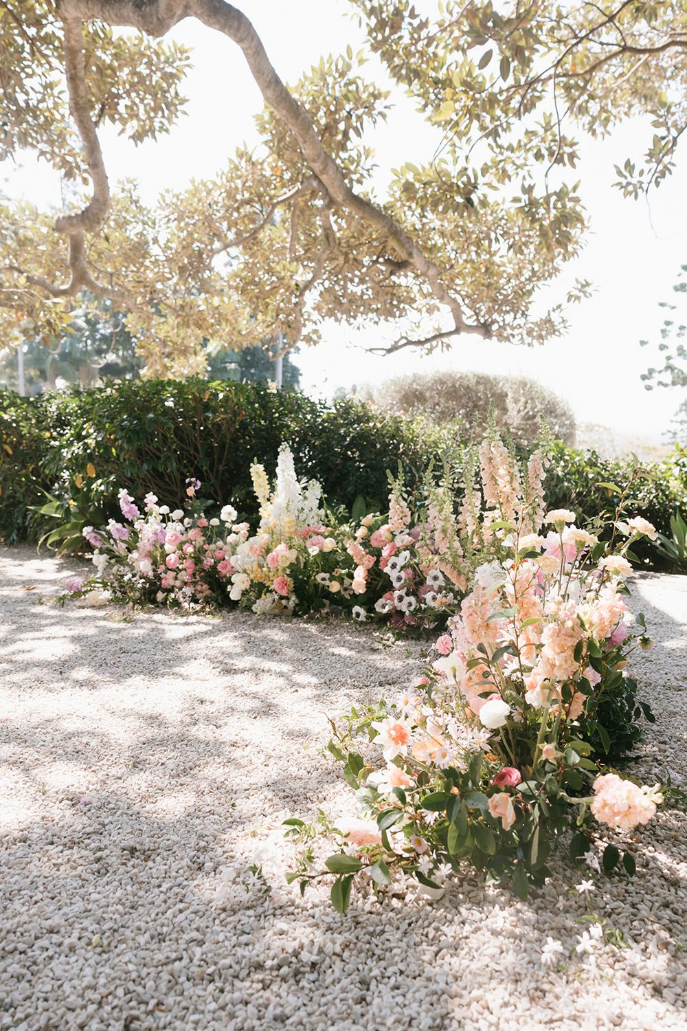 Beautiful outdoor garden ceremony setup with blush pink and white floral arrangements under a sunlit oak tree canopy.