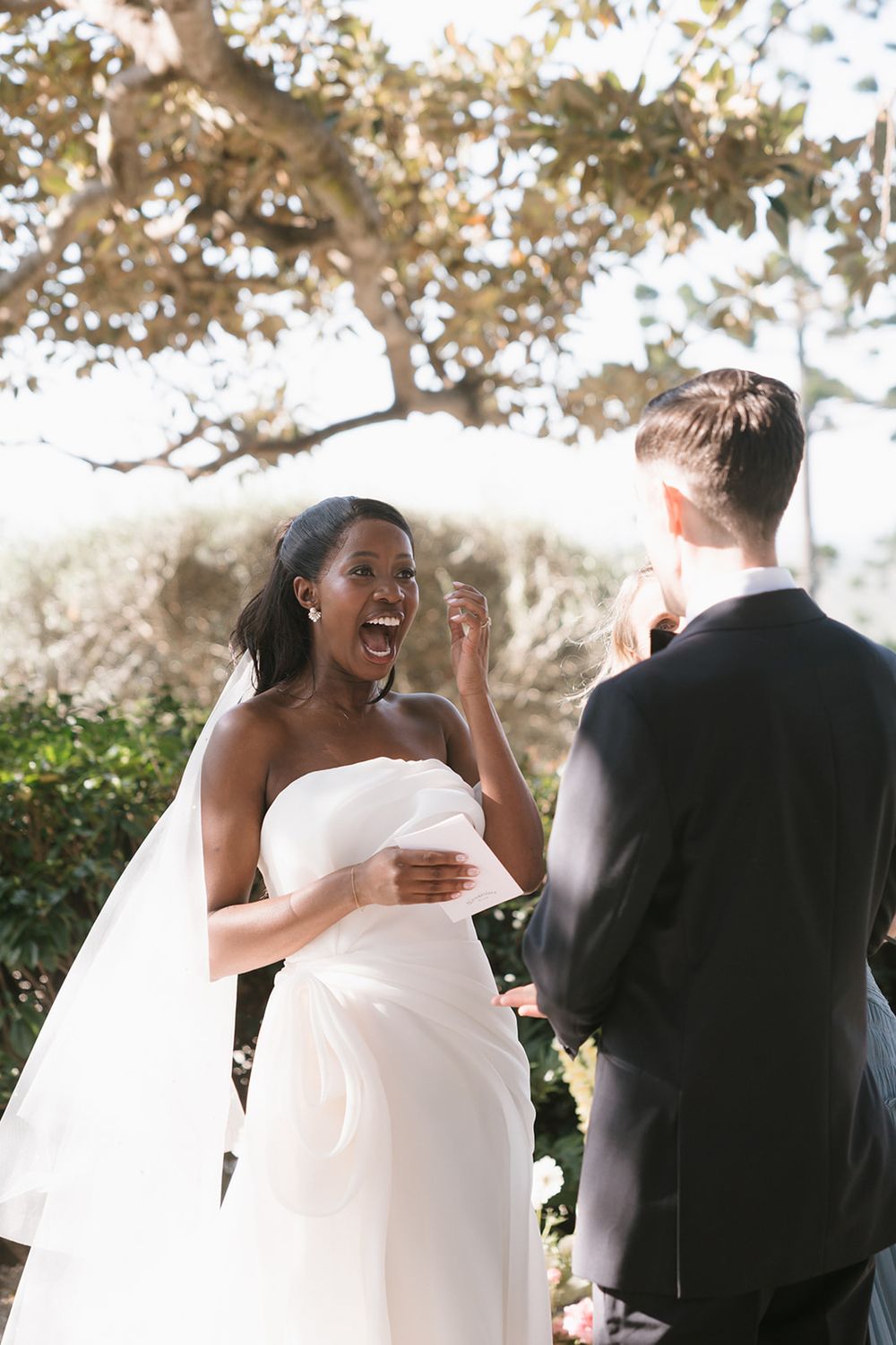 Joyful bride laughs with pure emotion during outdoor wedding ceremony beneath a tree.