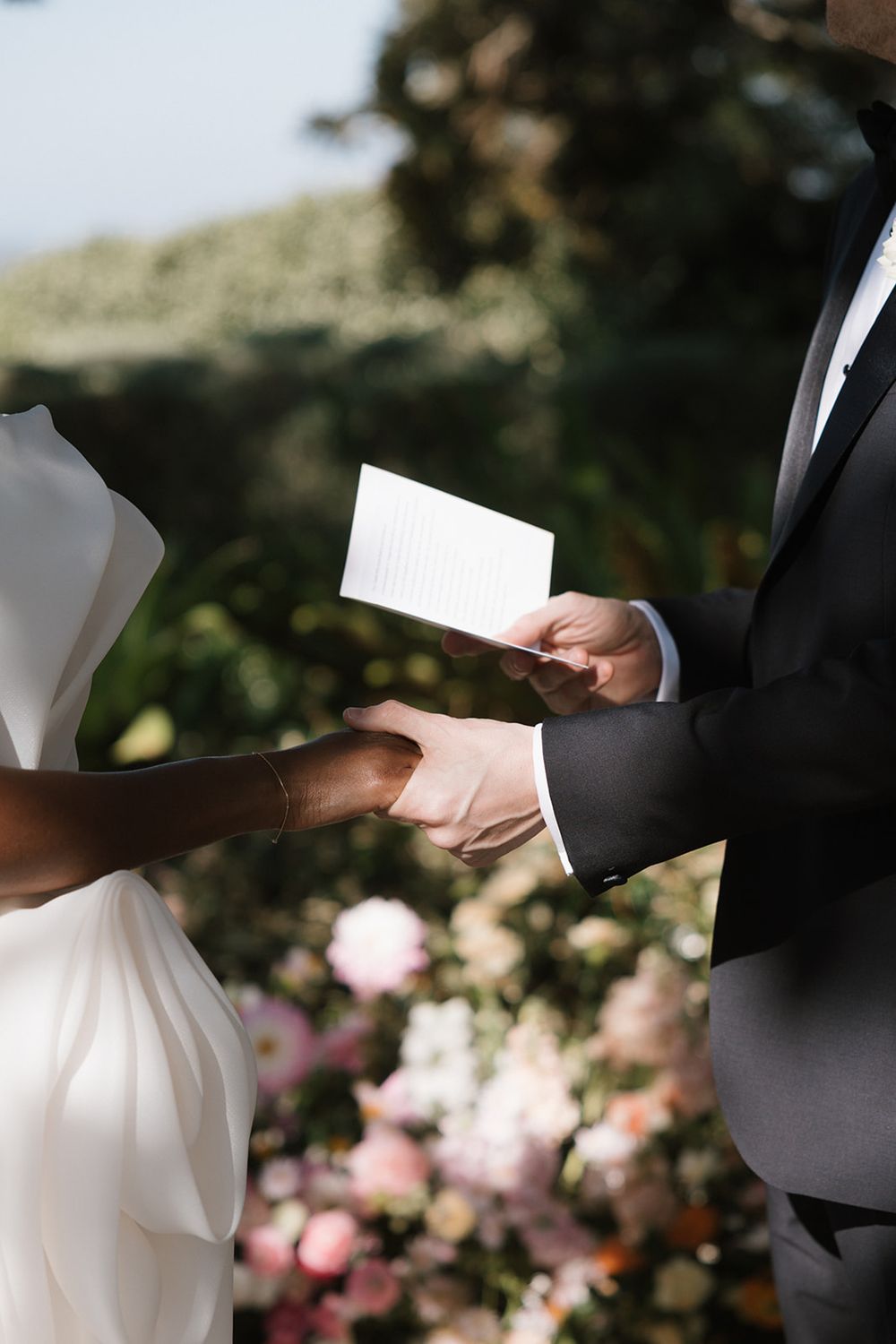 A couple holds hands during an outdoor wedding ceremony as the groom reads vows with pink roses in the background.