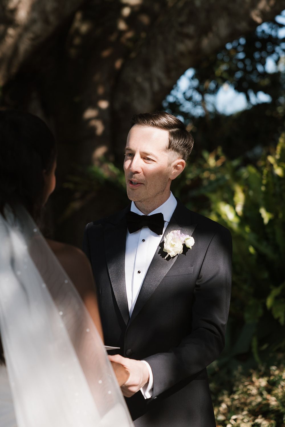 A groom in a black tuxedo with bowtie and white boutonniere smiles joyfully during an outdoor wedding ceremony.