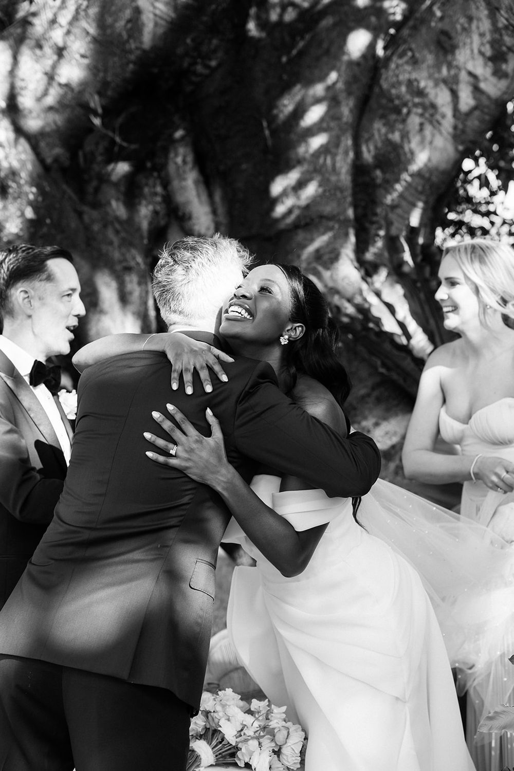 Wedding party members share joyous laughter and embraces during black and white outdoor celebration moment.
