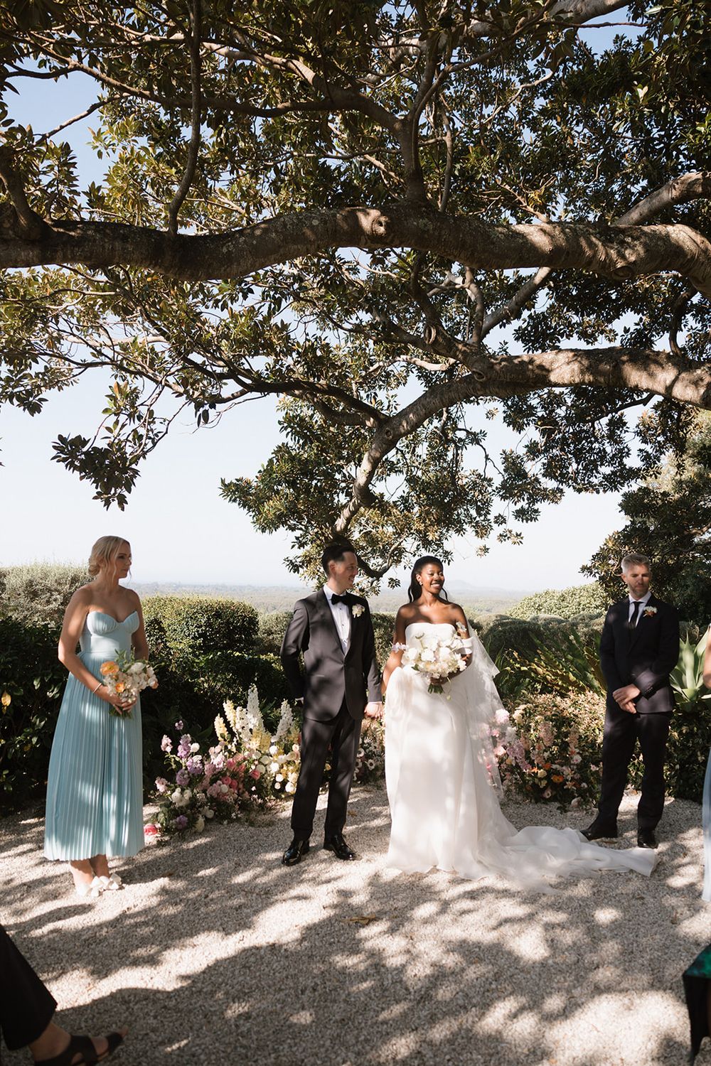 Wedding ceremony under a large tree canopy with bridesmaids and groomsmen during an outdoor celebration.