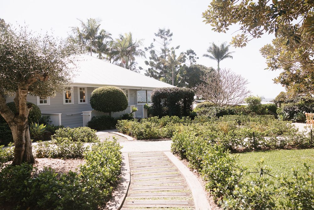 A curved stone pathway leads through manicured gardens to a white coastal home with palm trees and tropical landscaping.
