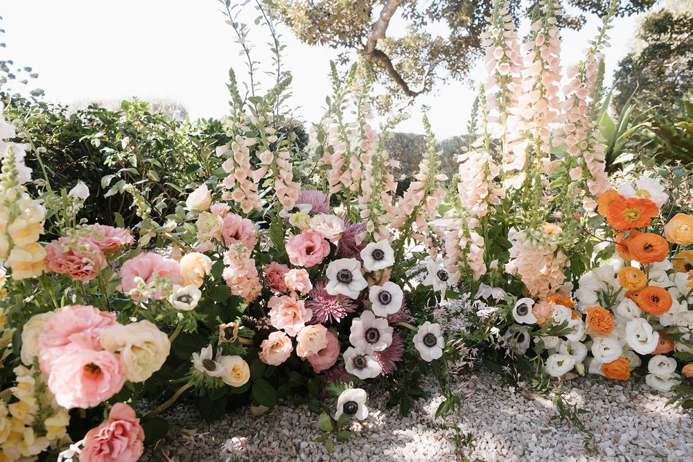 Beautiful garden-style floral arrangement with pink roses, anemones, and delphinium spikes in pastel spring colors.