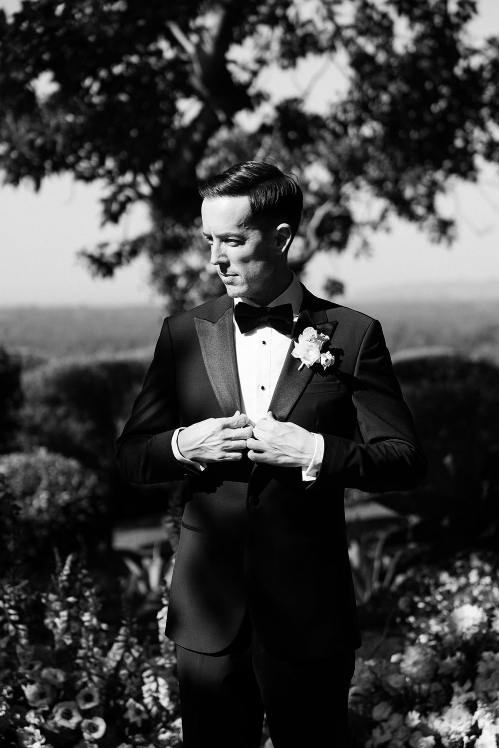 Black and white wedding photo of a groom in a tuxedo with boutonniere adjusting his cuffs in an outdoor garden setting.