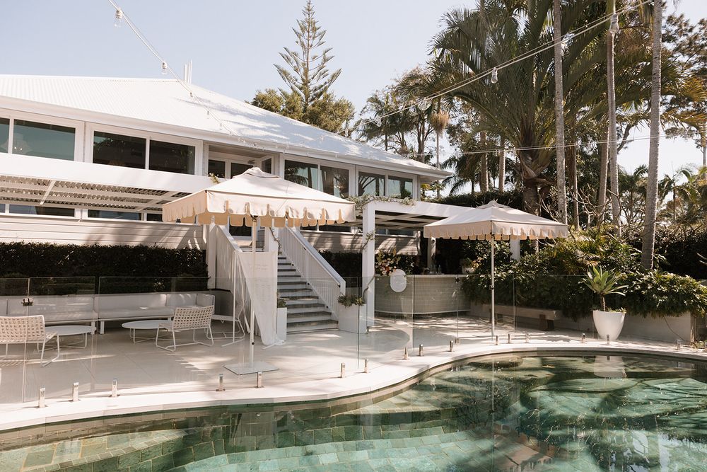 Modern white resort building with outdoor pool and lounge area featuring beige umbrellas and deck chairs.