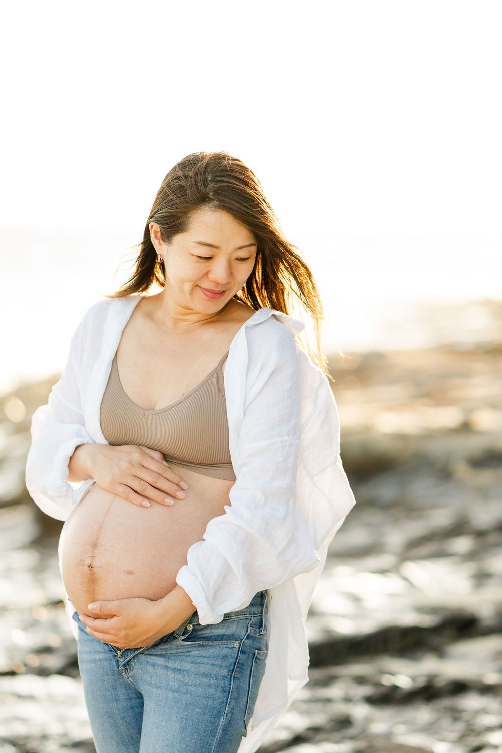 A pregnant woman in jeans and white cardigan stands on rocky terrain at golden hour with sunlight behind her.　Oahu Family Photographer | Aya