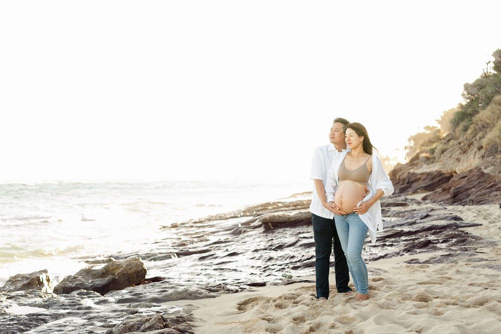 A couple embraces while standing on a rocky beach coastline during golden hour.　Oahu Family Photographer | Aya S Tomikawa Photography
