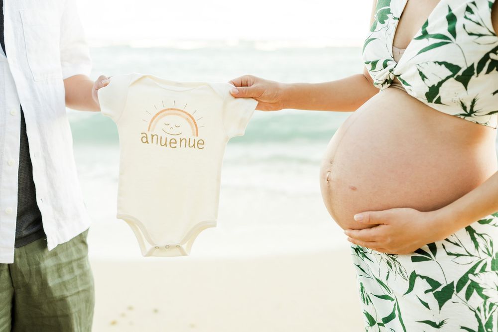 A pregnant woman in a floral dress being shown a small white baby onesie with orange text against a bright background.　Oahu Family Photograp