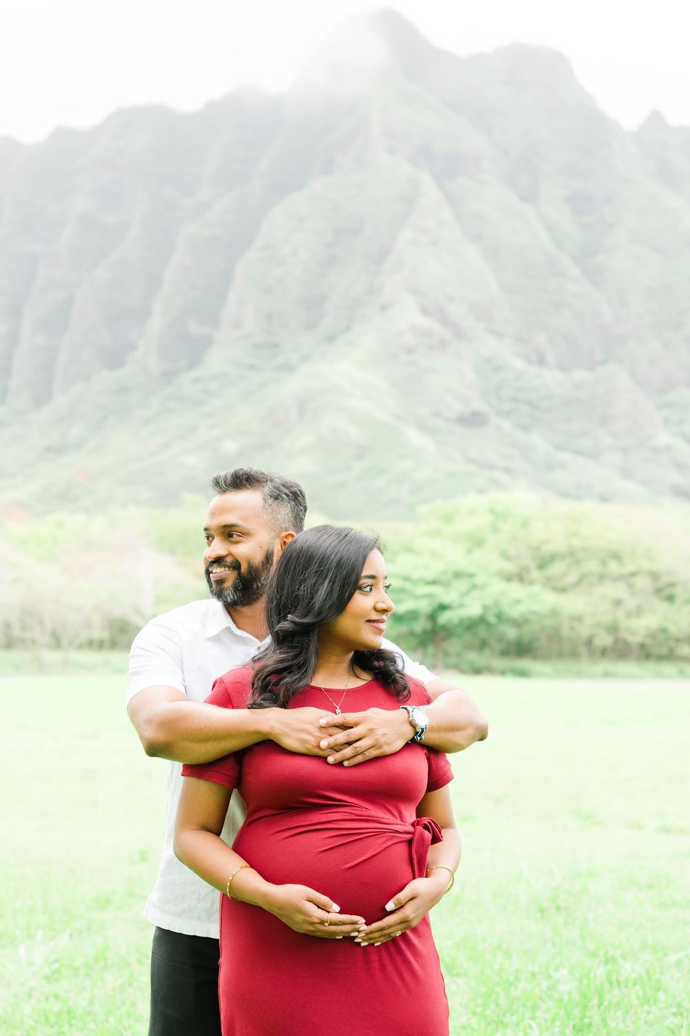 A couple embraces in front of majestic mountain peaks in Hawaii during a maternity photo session in nature.　Oahu Family Photographer | Aya S