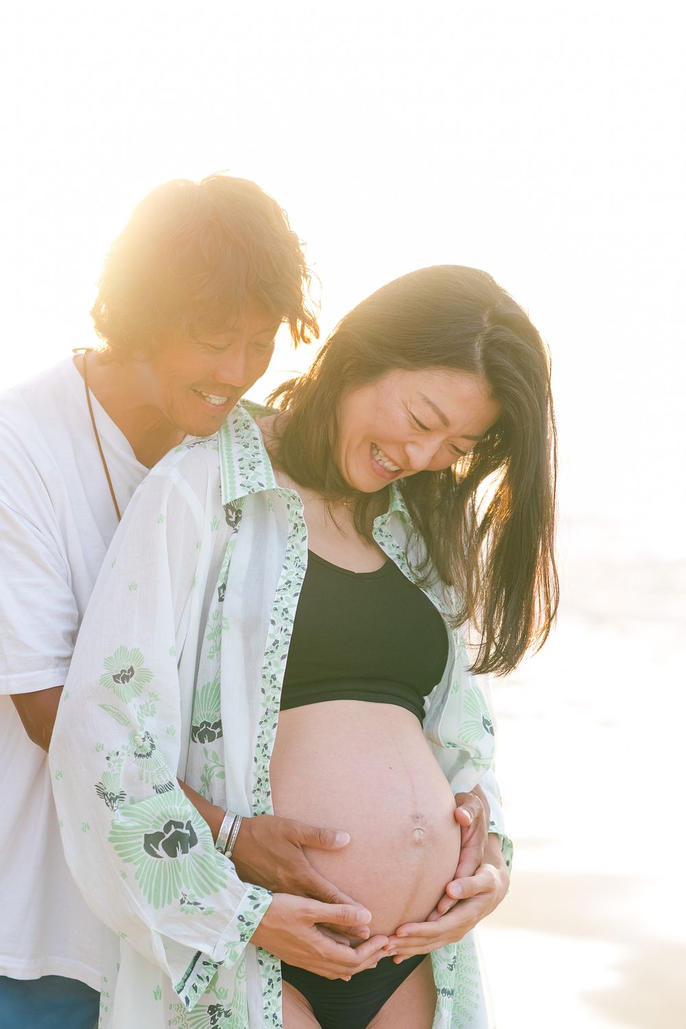 Expecting couple embraces on beach during sunset maternity photoshoot as woman wears mint green floral cover-upOahu maternity Photographer
