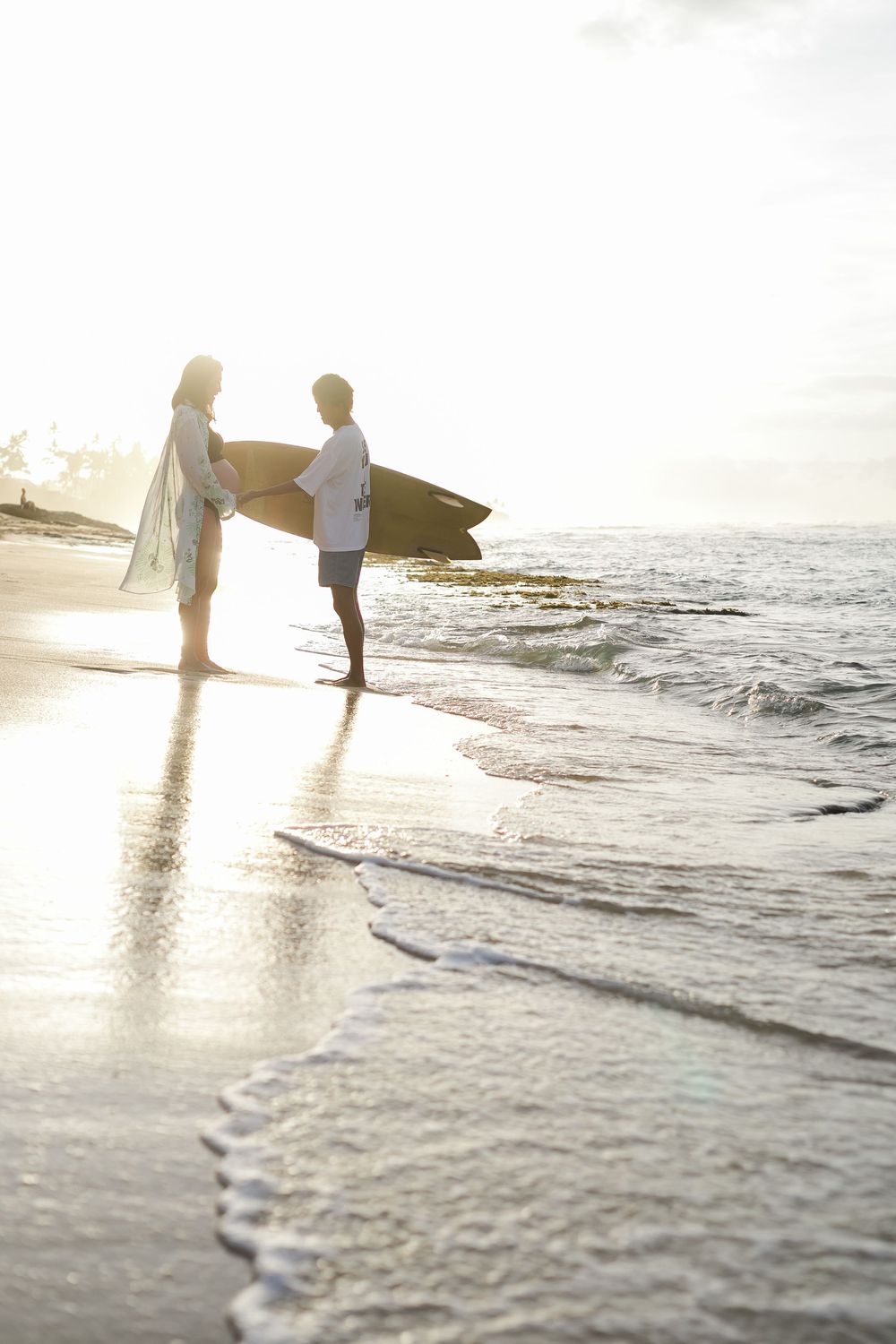 Two surfers stand silhouetted against a golden sunset on the beach as gentle waves roll onto the shore. Oahu maternity Photographer