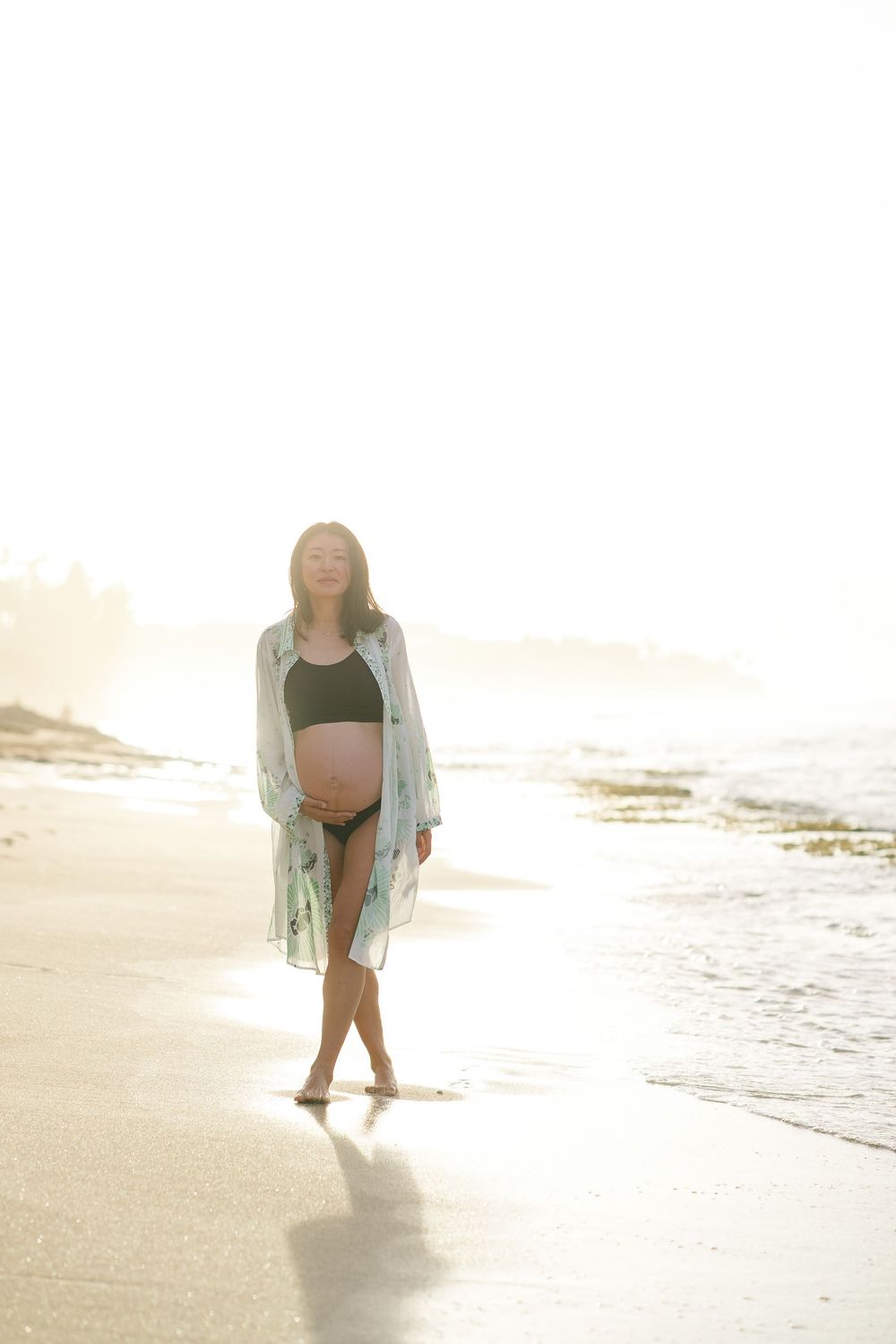 Silhouetted figure in a sheer coverup and swimwear walks along a sunlit beach at golden hour. Oahu maternity Photographer