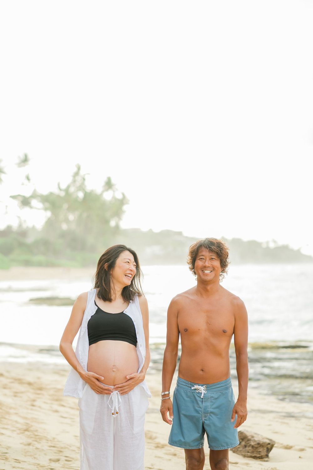 A happy couple sharing laughter together on a sunny beach, with waves and coastline in the background. Oahu maternity Photographer