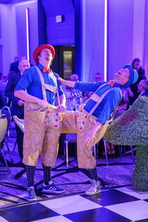 Two performers in lederhosen outfits dance energetically on a checkered floor under purple lighting at an event.