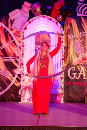A performer in red dress poses beneath an illuminated archway on stage with mystical carnival decorations.