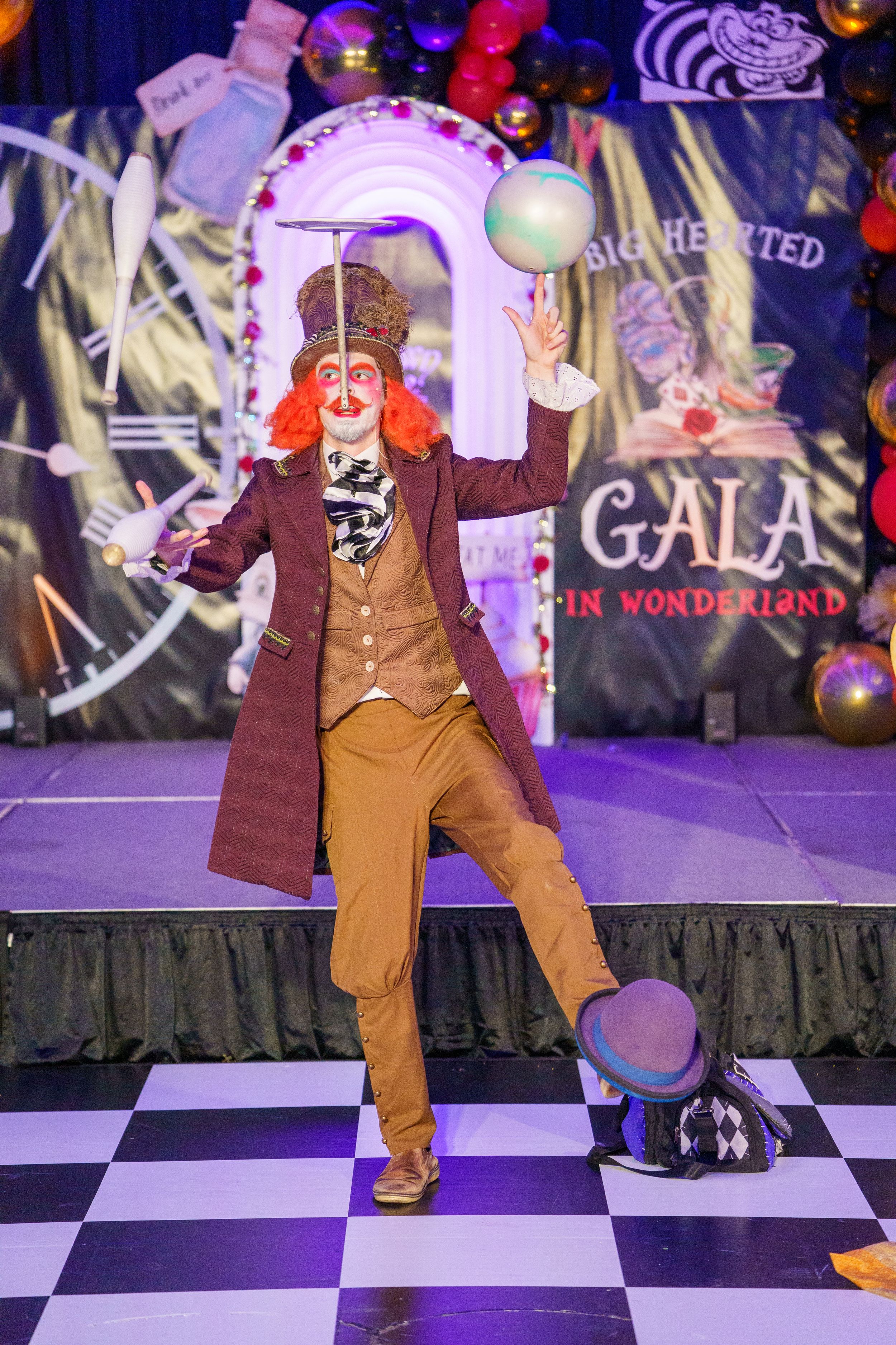A whimsical performer in a Victorian-style costume juggles on a checkered stage with colorful decorations and Gala signage.