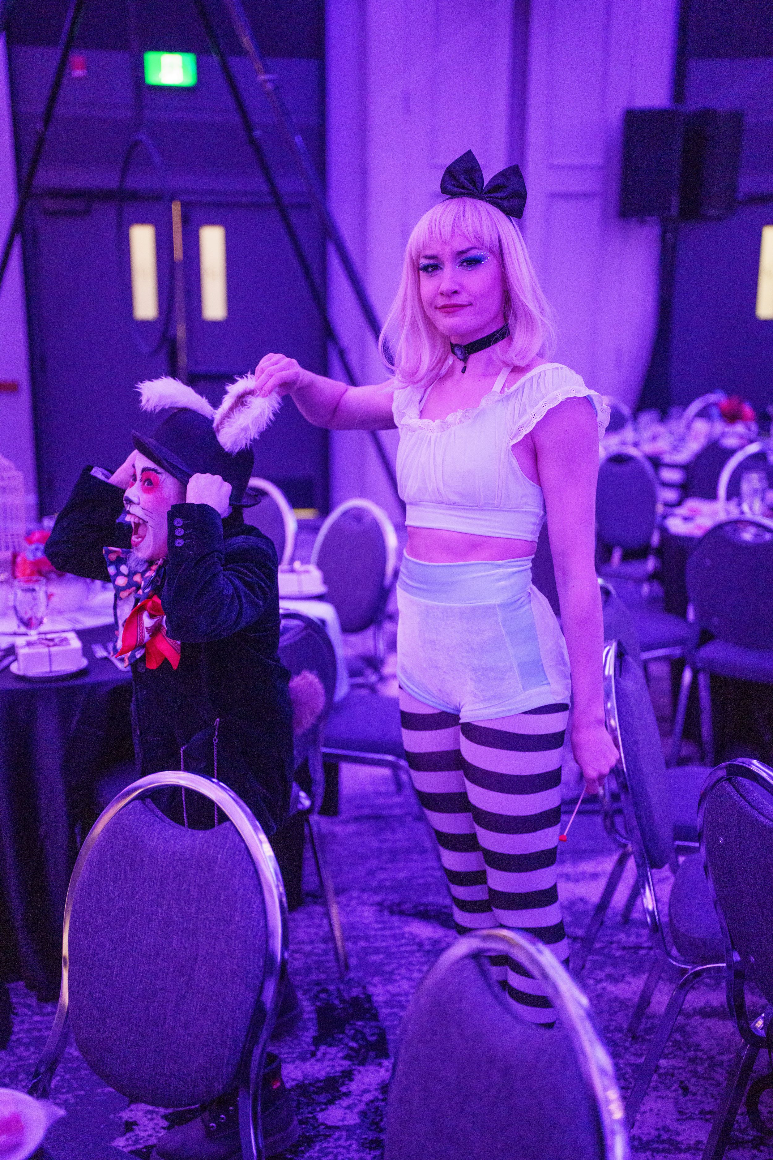 Whimsical purple-lit event space with striped leggings and black bow costume against backdrop of empty banquet chairs.