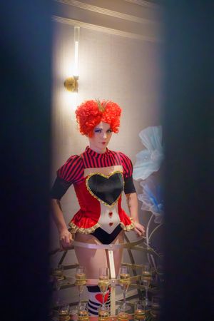 A costumed figure wearing a red and black corset outfit with striped socks and bright red curly wig stands in dramatic lighting.