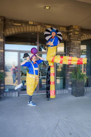 Two performers in blue and yellow costumes with umbrellas entertain outside a stone-accented building entrance.